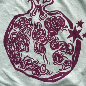 May include: A light mint green fabric featuring a dark purple screen-printed graphic of a halved pomegranate. The illustration displays the fruit's internal seeds and rind with bold, stylized lines, accompanied by two small four-pointed stars.