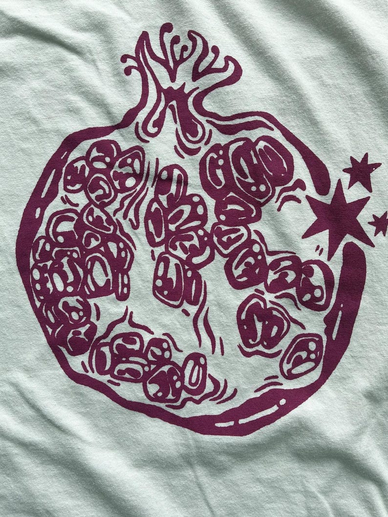 May include: A light mint green fabric featuring a dark purple screen-printed graphic of a halved pomegranate. The illustration displays the fruit's internal seeds and rind with bold, stylized lines, accompanied by two small four-pointed stars.