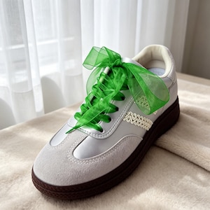 May include: A low-top sneaker featuring a smooth white upper with light gray suede overlays at the toe and heel. The shoe is laced with a wide, translucent green organza ribbon tied in a large bow. A decorative cream-colored braided trim accents the side, and the sneaker is finished with a dark brown textured rubber sole.