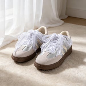 May include: A pair of low-top sneakers featuring a light beige suede toe cap, silver metallic side panels, and cream-colored textured stripes. The shoes are finished with white ruffled lace-style ribbon laces and a dark brown textured rubber sole.