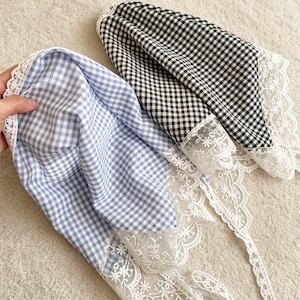 Può includere: Due fazzoletti triangolari con motivo a quadretti vichy, uno azzurro e bianco e l'altro nero e bianco. Ogni fazzoletto è rifinito con un ampio e intricato bordo in pizzo floreale bianco lungo i lati ed è dotato di lunghi lacci.
