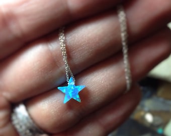 May include: A delicate necklace featuring a small, five-pointed star pendant. The star is a vibrant blue with a shimmering, opal-like effect. The chain is a thin, silver-colored metal, creating a simple and elegant design.