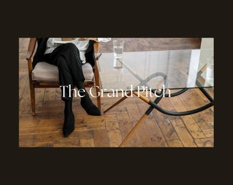 May include: A person seated in a wooden chair, wearing black pants and shoes, with the text "The Grand Pitch" overlaid. A glass table with a curved black and wood base is in the foreground, and a glass of water is on the table.
