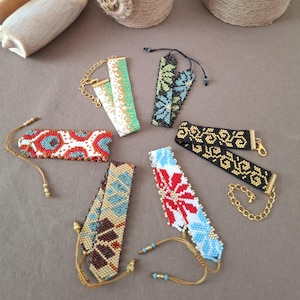 Puede incluir: Una colección de pulseras de cuentas en varios colores y diseños, incluyendo rojo, azul, verde y dorado. Las pulseras están dispuestas alrededor de un jarrón decorativo envuelto en cuerda, una concha y una pequeña caja redonda.