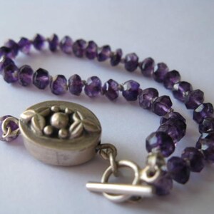 May include: A purple amethyst gemstone bracelet with a silver clasp featuring a floral design. The bracelet is made of faceted beads and has a toggle clasp.