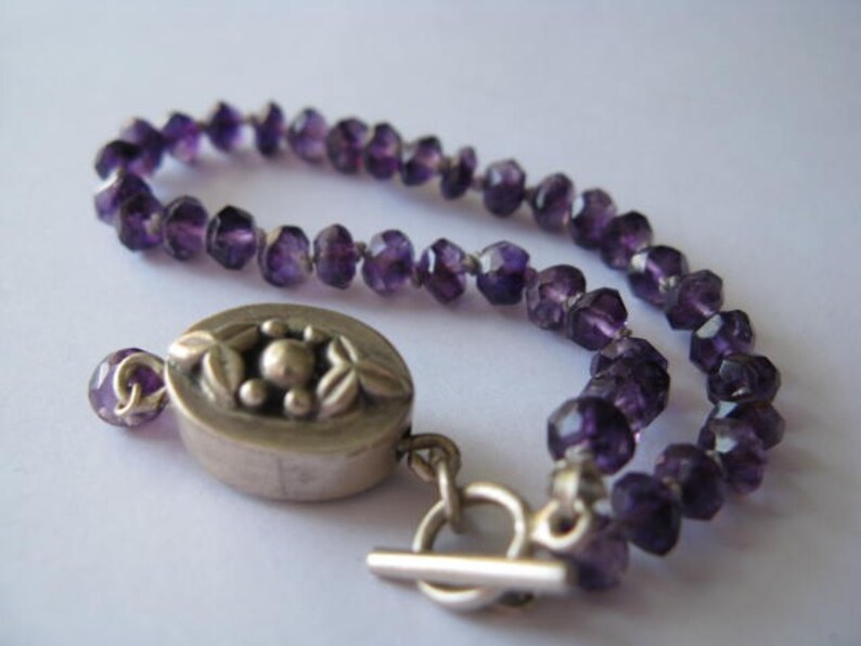 May include: A purple amethyst gemstone bracelet with a silver clasp featuring a floral design. The bracelet is made of faceted beads and has a toggle clasp.