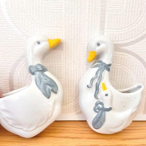 May include: Two white ceramic wall-mounted planters shaped like geese. Each goose features a yellow beak and a grey painted bow around its neck. The right planter includes a smaller goose figure attached to the larger one, also wearing a grey bow.