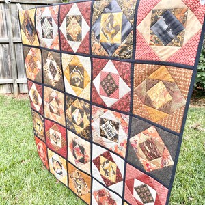 May include: A handmade quilt with a patchwork design. The quilt features a grid of squares, each containing a diamond-shaped pattern in shades of brown, red, and gold. The quilt is hanging outside on a washing line.