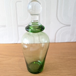 May include: A clear glass bottle with a tapered body and a rounded base, featuring a distinct green tint that deepens towards the bottom. The bottle includes a matching green glass collar and a clear, spherical glass stopper at the top.