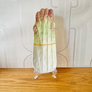 May include: White ceramic asparagus dish with green and purple painted details. The dish is shaped like a bunch of asparagus and has a yellow painted ribbon around the middle.