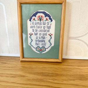 May include: Framed cross-stitch artwork in a wooden frame. The artwork features a quote in blue thread on a light green background, with floral and heart motifs. The quote reads: "A woman has to work twice as hard to be considered half as good as a man - fortunately that's easy!"