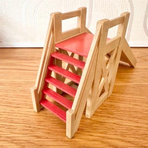 May include: A miniature wooden toy slide featuring a natural light-colored wood frame and bright red steps. The structure includes a small platform at the top of the stairs and a smooth, red-painted sliding surface extending downward to the base.