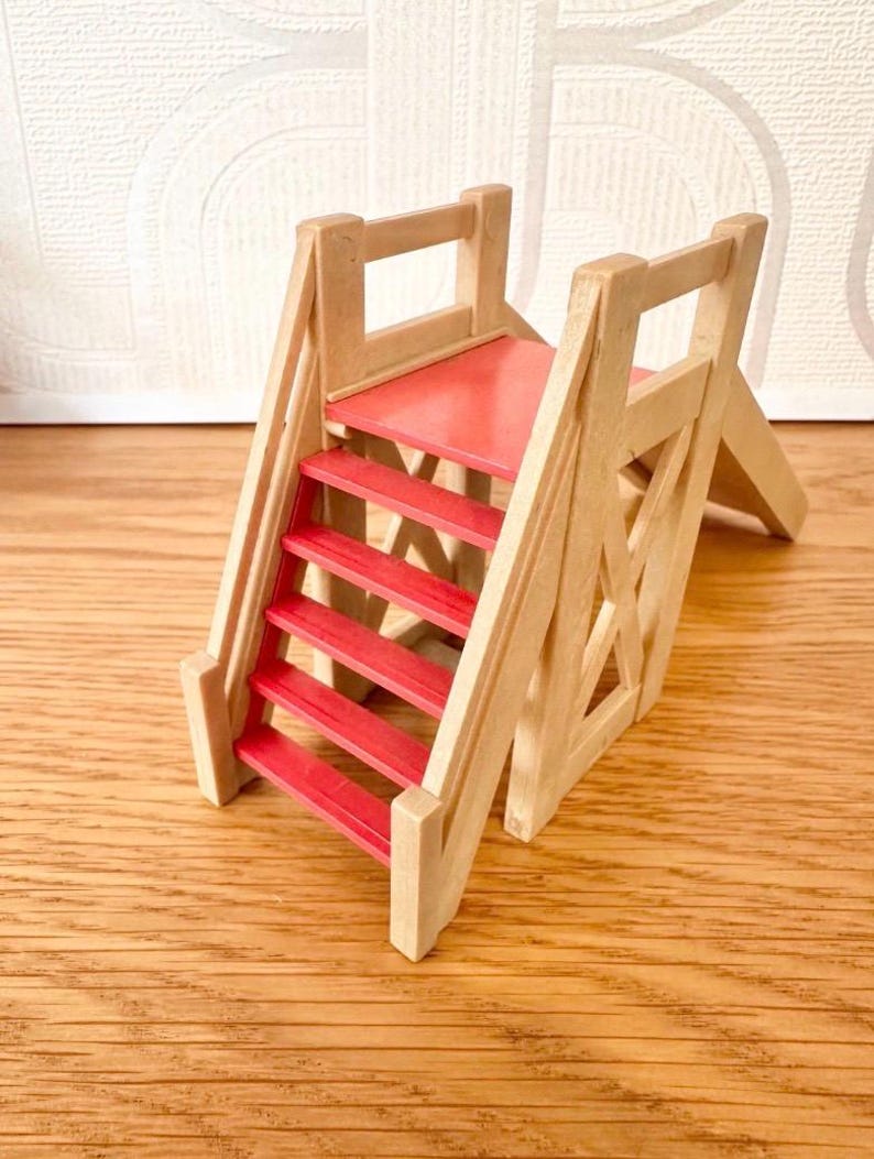 May include: A miniature wooden toy slide featuring a natural light-colored wood frame and bright red steps. The structure includes a small platform at the top of the stairs and a smooth, red-painted sliding surface extending downward to the base.