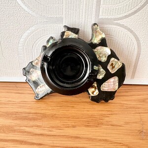 May include: Black ashtray shaped like Australia, decorated with iridescent shell fragments. The ashtray features a circular recess for holding ashes and cigarettes. It is placed on a wooden surface.