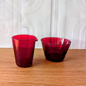 May include: A set of two ruby red glass containers, one a small jug and the other a bowl. The containers are on a wooden surface.