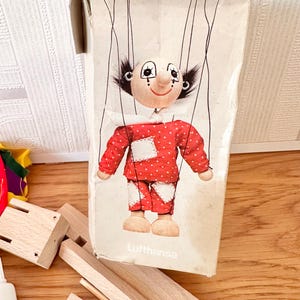 May include: A vintage puppet in its original packaging. The puppet has a painted face with black hair and is dressed in a red outfit with white polka dots and patches. The packaging is a cardboard box with the word "Lufthansa" printed on it.