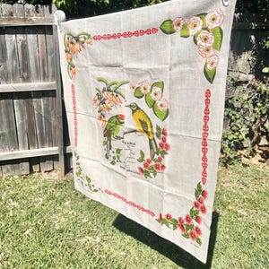 May include: A beige linen tea towel featuring a central illustration of a yellow and green bird, a map of New Zealand, and the text Aotearoa New Zealand. The border is decorated with red geometric patterns and floral motifs in the corners.