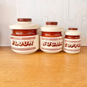 May include: A trio of ceramic kitchen storage jars with lids. The jars are off-white with a reddish-brown band and matching lids. The words "FLOUR", "SUGAR", and "COFFEE" are printed on the front in reddish-brown.