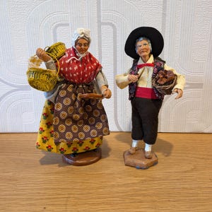May include: Two decorative figurines depicting an elderly pair in traditional attire. The figure on the left wears a white cap, red patterned shawl, and brown apron over a yellow floral skirt, carrying two yellow baskets. The figure on the right wears a black hat, dark vest, and brown trousers, holding a small basket.