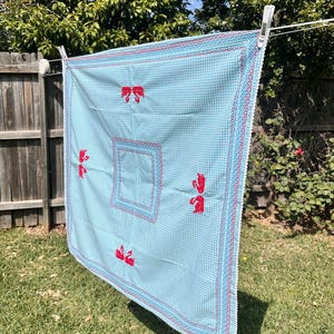 May include: A square light blue and white gingham fabric cloth featuring red cross-stitched swan motifs arranged around a central square border. The edges are finished with a decorative white scalloped trim and a red zigzag stitched pattern.