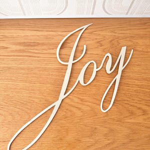 May include: A cream-colored wooden sign with the word "Joy" in a cursive font. The sign is placed on a wooden surface with a light wood grain pattern. The background is a white patterned wall.
