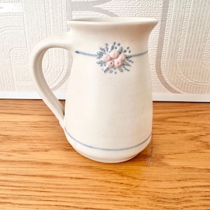 May include: A cream-coloured ceramic pitcher with a single handle, featuring a hand-painted blue horizontal line and a small, raised floral motif in pink and blue near the rim. The pitcher has a smooth, matte finish and a simple, rounded shape.