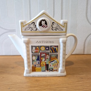 May include: A white ceramic teapot shaped like a building with a gabled roof lid. The front features a colourful illustration of an antique shop storefront with the text "ANTIQUES" above and "an antique teapot" printed along the bottom edge.