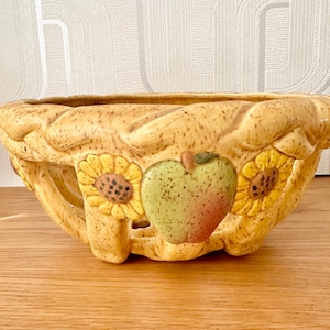 May include: A ceramic fruit bowl with a woven design and a speckled glaze. The bowl features three sunflowers and an apple in the centre.