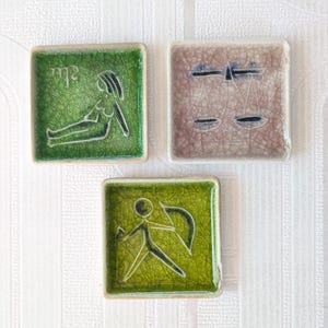 May include: Three square ceramic tiles with zodiac symbols. One green tile features a reclining figure, another shows scales, and the third depicts an archer. The tiles have a textured, crackled finish and are set against a white background.