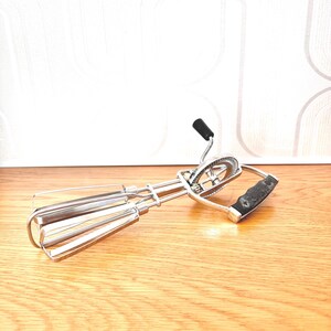 May include: A manual rotary egg beater constructed from polished stainless steel. The kitchen tool features two wire beaters, a central gear mechanism with a small black handle, and a larger black textured handle for a secure grip during use.