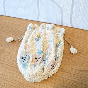 以下が含まれることがあります： 白のシードビーズで覆われた小さな巾着袋で、水色、ピンク、黄色の花びらの模様が施されています。バッグの開口部はギャザーが寄っており、両サイドにビーズ付きの引き紐が2本付いていて、口を閉じることができます。