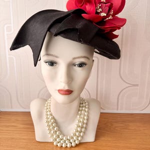 May include: A black hat with a large red flower and white detailing. The hat is displayed on a mannequin head, paired with a triple-strand pearl necklace. The hat has a distinctive, layered design.