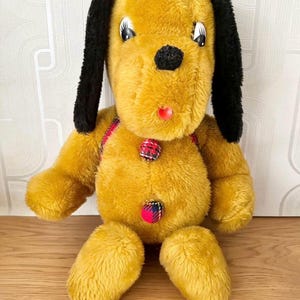 May include: A plush yellow dog toy with black ears and a red tongue sticking out. The dog has a plaid pattern on its chest and three buttons.