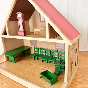 May include: A wooden dollhouse with a pink roof and two levels. The top floor contains a red grandfather clock and a small bench. The bottom floor features a long green table, four green chairs, a green high chair, and a small green cradle.