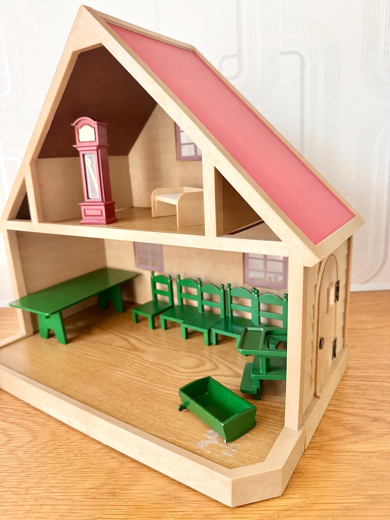 May include: A wooden dollhouse with a pink roof and two levels. The top floor contains a red grandfather clock and a small bench. The bottom floor features a long green table, four green chairs, a green high chair, and a small green cradle.