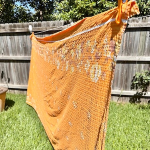 May include: An orange textile with a floral pattern is hanging on a washing line with an orange peg. The fabric is draped over a washing line, with a wooden fence and green grass in the background. The textile has a white stripe.