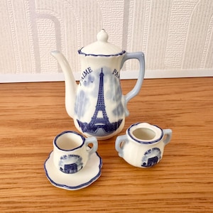 May include: A miniature porcelain tea set in white and blue. The set includes a teapot with an Eiffel Tower design, a teacup with a saucer, and a sugar bowl. The teapot has the words "J'AIME PARIS" on it. The set is a charming souvenir.