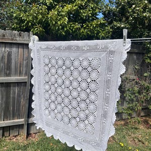 May include: A square white crochet blanket featuring a repeating floral motif pattern in the centre, surrounded by a wide decorative border with intricate stitching and a scalloped edge, hanging from a washing line by two metal clips.