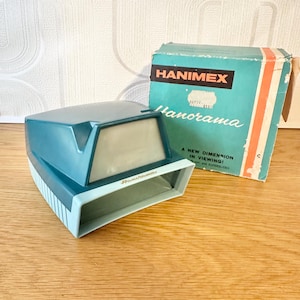 May include: A teal and light blue Hanimex Panorama slide viewer with its original box. The box displays the Hanimex logo, the word "Panorama", and the slogan "A New Dimension in Viewing!" printed on it.