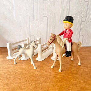 May include: A toy equestrian set featuring a rider in a red jacket and black helmet on a tan horse. A white foal stands nearby, with a white fence in the background. The set is on a wooden surface.