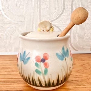 May include: A cream-coloured ceramic honey pot with a matching lid featuring a small bee figurine. The pot is decorated with painted blue and orange flowers and green grass. A wooden honey dipper is inserted through the lid opening.