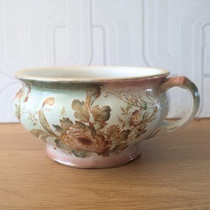 May include: An antique ceramic bowl with a floral pattern. The bowl is painted in light green and pink, with brown and gold floral details. It has a handle and a wide rim, and is set on a small base.