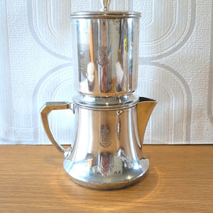 以下が含まれることがあります： 円筒形のトップと注ぎ口が付いた、ヴィンテージの銀色のコーヒーポット。 ポットにはハンドルと小さなノブが付いた蓋が付いています。表面は周囲を反射しています。 コーヒーポットは木製の表面に置かれています。