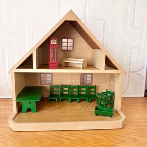 May include: A two-story wooden dollhouse with a triangular roof. The top floor contains a red grandfather clock and a beige bench. The ground floor features a green table, four green chairs, a green high chair, and a small green crate.
