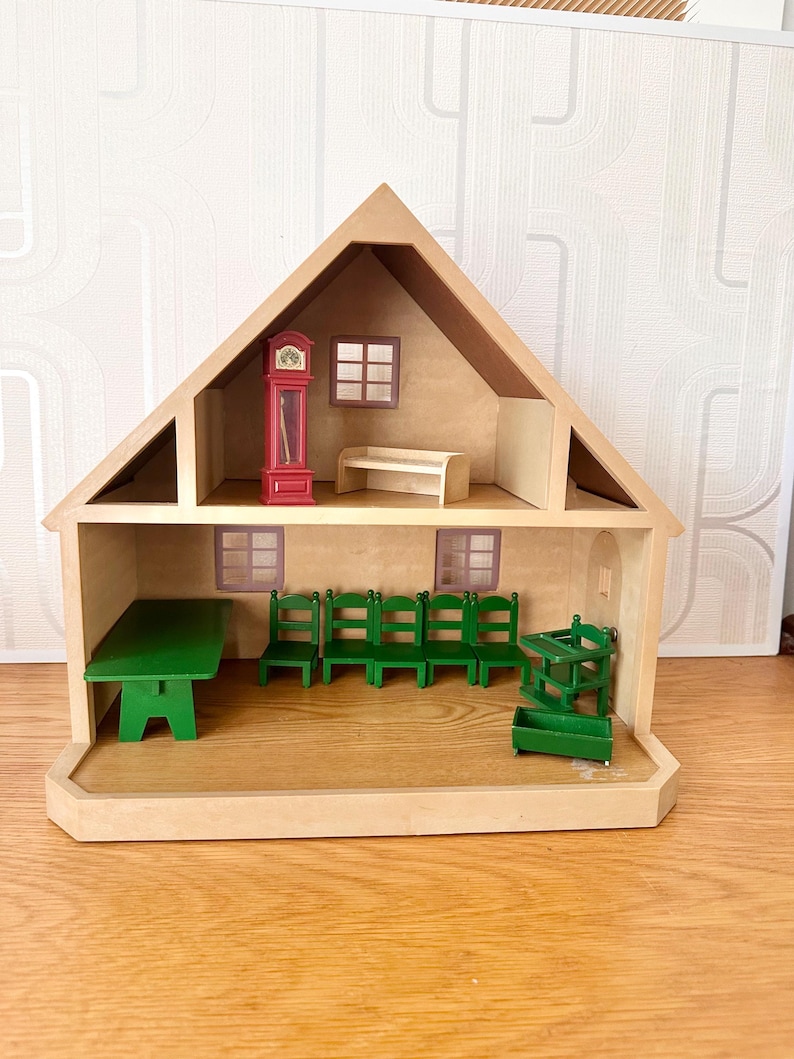 May include: A two-story wooden dollhouse with a triangular roof. The top floor contains a red grandfather clock and a beige bench. The ground floor features a green table, four green chairs, a green high chair, and a small green crate.