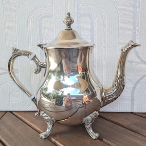 May include: A silver teapot with a lid and ornate handle and spout. The teapot sits on three decorative feet. The polished surface reflects the surroundings. The teapot is likely made of silver or a silver-colored metal.