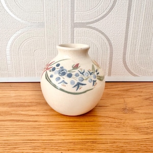 May include: A small, round, off-white ceramic vase featuring a hand-painted botanical design. The decoration includes clusters of blue berries, green leaves, and small red flower buds encircling the upper portion of the bulbous body.