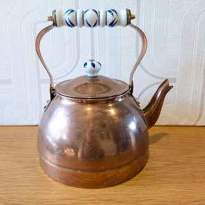 May include: A polished copper tea kettle with a rounded body and a curved spout. The handle features a white ceramic grip with blue leaf-like patterns, and the lid is topped with a matching white and blue ceramic knob with a metal base.