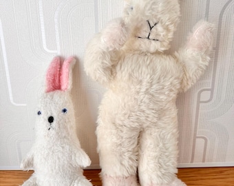 May include: Two plush rabbit toys with white fur and pink inner ears. The smaller rabbit has blue eyes and a black nose. The larger rabbit has embroidered facial features, including a Y-shaped nose and mouth, and pink accents on its feet.