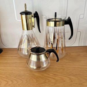 May include: Three glass coffee carafes with gold accents and black lids. The carafes have different patterns, including a geometric pattern and a leaf pattern.